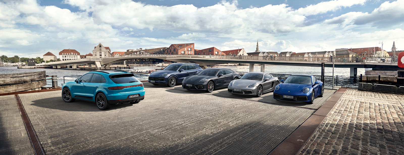 Mehrere Porsche Sportwagen stehen auf einem Parkplatz nahe einer Brücke.